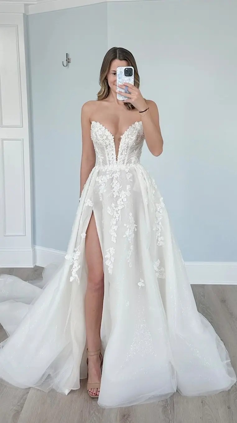Model is wearing an a-line strapless wedding dress with a plunging neckline and thigh high slit