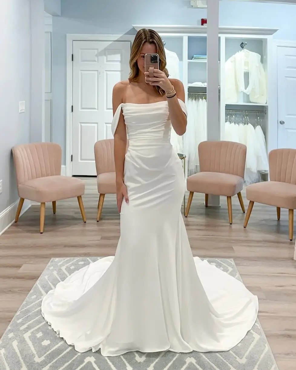 Model is wearing an off the shoulder minimalist bridal gown with a fit and flare silhouette and ruching on the bodice