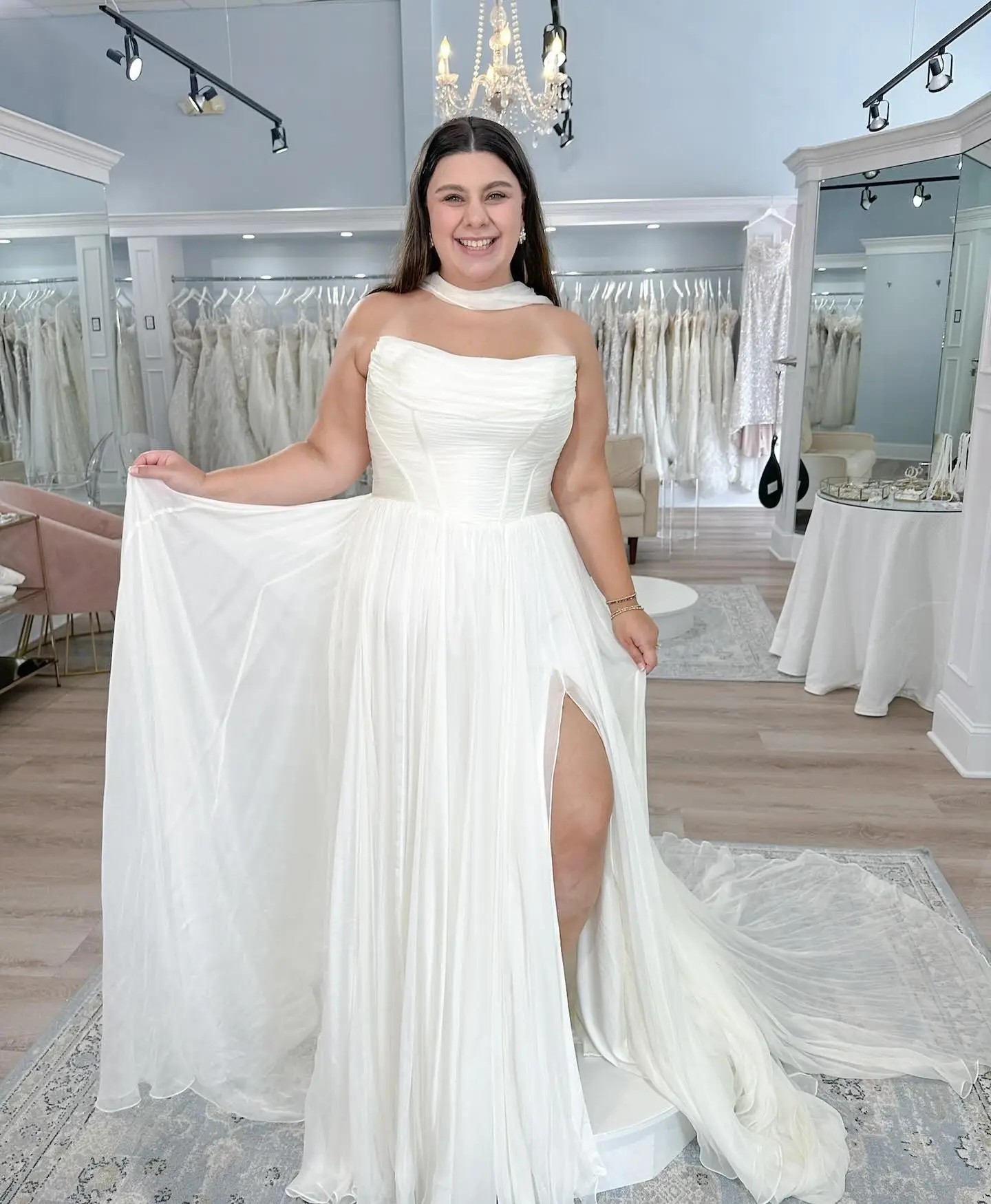 Model is wearing a strapless a-line wedding gown with a scoop neckline and pleated skirt with a high slit and bridal scarf